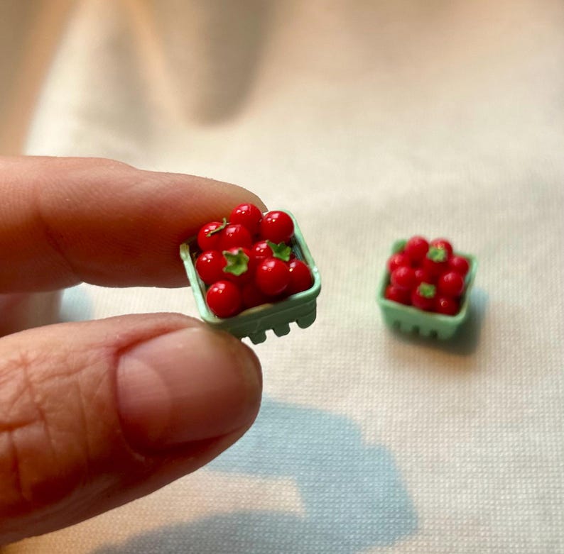 May include: Miniature red tomatoes with green stems are displayed in a small, light green berry basket. The basket is held in a person's hand, with another basket of tomatoes in the background. The tomatoes are a vibrant red color.