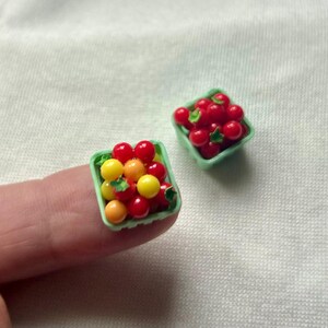May include: Miniature red, yellow, and orange tomatoes in small green plastic baskets. The tomatoes are arranged to look like they were just picked. The baskets are sitting on a white surface.