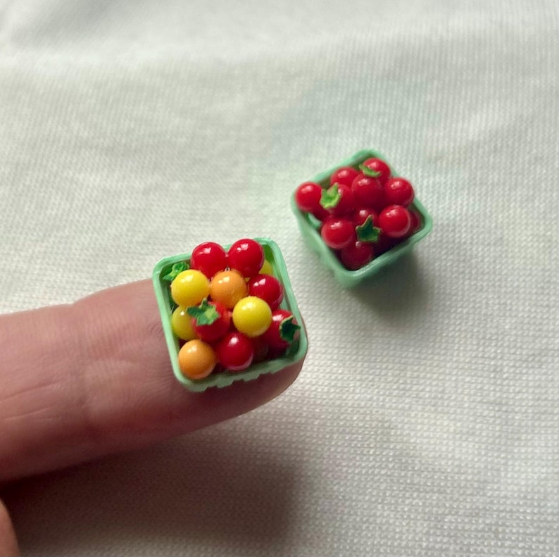 May include: Miniature red, yellow, and orange tomatoes in small green plastic baskets. The tomatoes are arranged to look like they were just picked. The baskets are sitting on a white surface.
