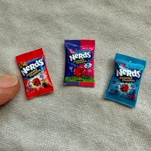 May include: Three miniature bags of Nerds Gummy Clusters candy in red, blue, and purple. The bags have the Nerds logo and images of the candy. The bags are on a gray textured surface.