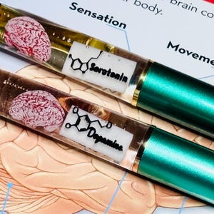 May include: Two pens with clear barrels, each containing a miniature brain model. One pen has a label that says "Serotonin" and the other says "Dopamine". The pens have green metallic barrels and gold accents. The background includes a diagram of the brain.