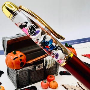 May include: A burgundy and gold pen with a clear barrel filled with Halloween-themed charms. The image also features miniature pumpkins, wooden leaves, and toy knives, creating a festive seasonal display.