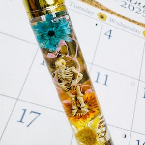 May include: A unique pen with a gold-colored top and black base. The clear barrel encases a skeleton figurine, blue and yellow flowers, and pink accents. The pen is a decorative writing instrument.