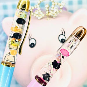 May include: Two pens with clear barrels filled with small, colorful charms. One pen has a blue barrel and gold accents, the other has a pink barrel and gold accents. The background features a pink piggy bank and a jeweled crown.