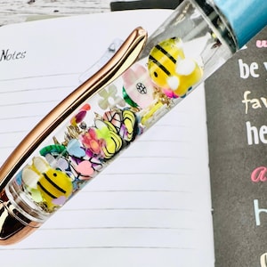 May include: A clear pen filled with colourful bee and flower-shaped charms, gold flakes, and a light blue top. The pen has a rose gold clip and tip, resting on a lined notebook page with the word "Notes".