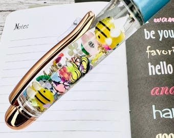 May include: A clear pen filled with colorful bee and flower-shaped charms, gold flakes, and a light blue top. The pen has a rose gold clip and tip, resting on a lined notebook page with the word "Notes".