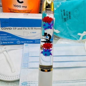 May include: A white pen with a gold cap and a clear barrel containing red and blue virus-shaped figures and a black microscope. The pen is surrounded by medical supplies, including a COVID-19 test kit, face masks, and a vitamin C supplement.
