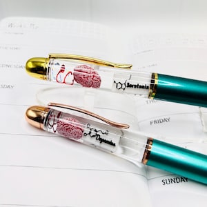 Puede incluir: Dos bol&iacute;grafos con barriles transparentes, detalles dorados y oro rosa, y cuerpos verde azulado. Los barriles contienen un modelo de cerebro rojo y las palabras "Serotonina" y "Dopamina". Los bol&iacute;grafos est&aacute;n sobre un planificador.