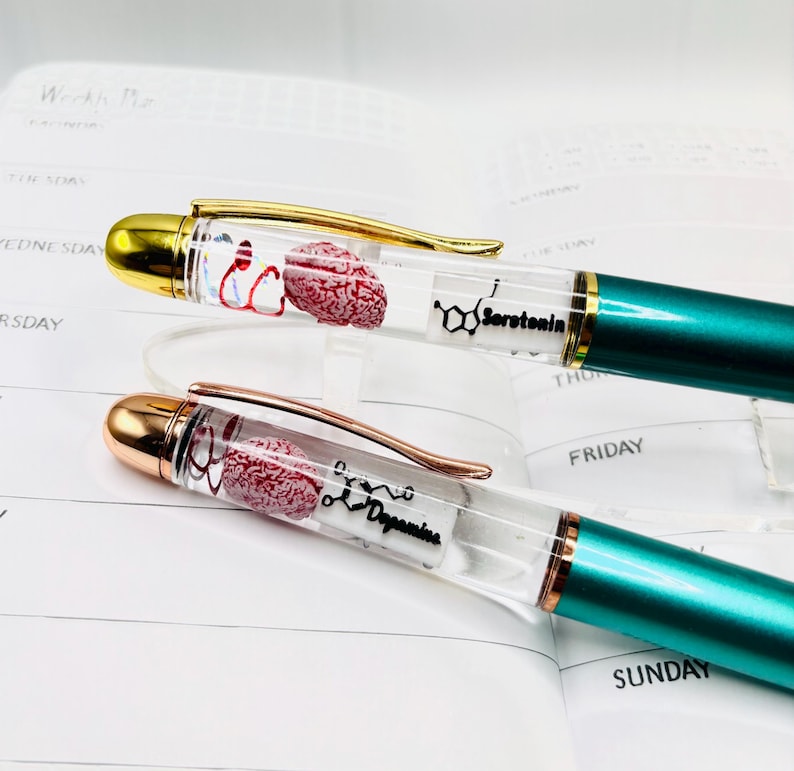 Puede incluir: Dos bol&iacute;grafos con barriles transparentes, detalles dorados y oro rosa, y cuerpos verde azulado. Los barriles contienen un modelo de cerebro rojo y las palabras "Serotonina" y "Dopamina". Los bol&iacute;grafos est&aacute;n sobre un planificador.