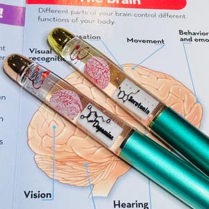 Puede incluir: Dos bol&iacute;grafos con barriles transparentes, tapas doradas y bases verde azulado. Los barriles contienen un cerebro rosa en miniatura y una etiqueta blanca con la palabra "Serotonina" y una estructura qu&iacute;mica. Los bol&iacute;grafos descansan sobre un libro abierto con un diagrama del cerebro.