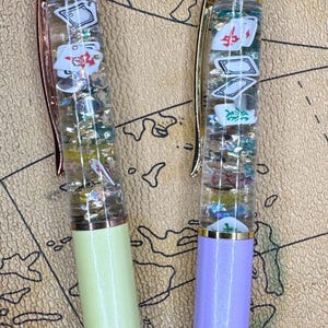 May include: Two novelty pens with clear barrels filled with floating playing card-themed charms and glitter. One pen has a light green base and rose gold accents, the other has a purple base and gold accents. Both have a map background.