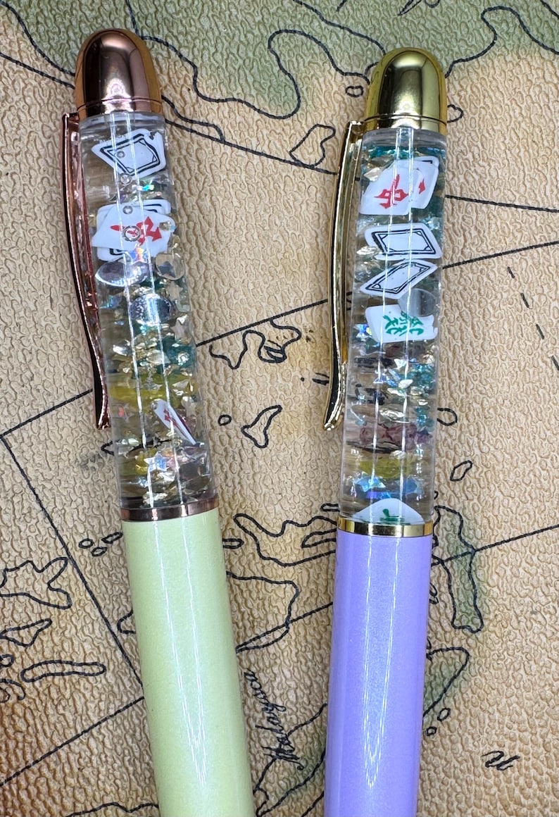May include: Two novelty pens with clear barrels filled with floating playing card-themed charms and glitter. One pen has a light green base and rose gold accents, the other has a purple base and gold accents. Both have a map background.