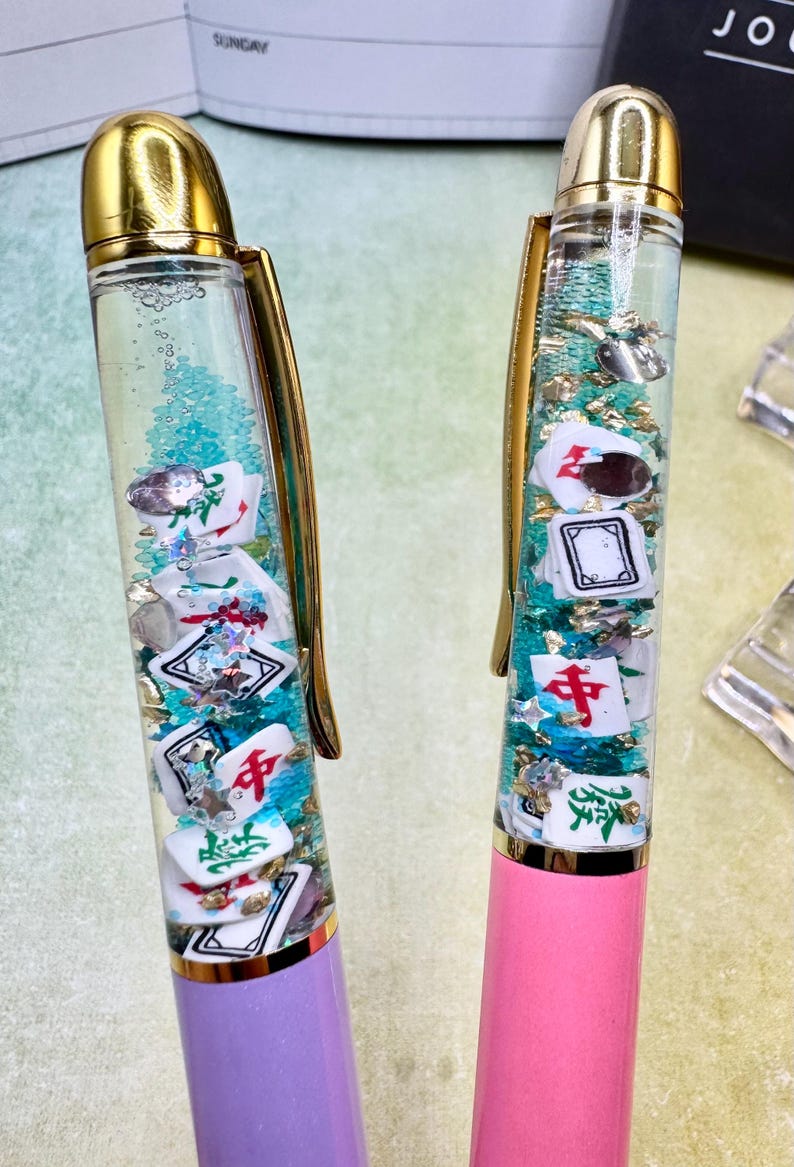 May include: Two pens with gold accents, one lavender and one pink. The clear barrels are filled with water, glitter, and miniature Mahjong tiles. The pens have gold clips and tops.