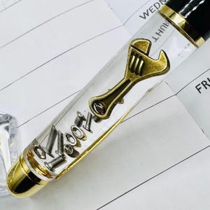 May include: A close-up of a pen with a clear barrel filled with small metal bolts and a gold-colored wrench charm. The pen has a black top and gold accents. The pen is resting on a white paper with lines.