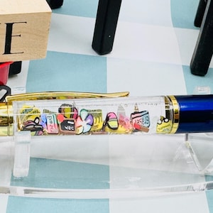 May include: A blue and gold pen with a clear barrel filled with colourful miniature objects. The pen has a gold clip and tip, and the barrel is filled with small, decorative items. The pen is displayed on a clear stand.