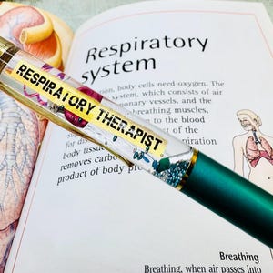 May include: A green and gold pen with the words "RESPIRATORY THERAPIST" on a yellow label. The pen is lying on an open book with illustrations of the respiratory system.