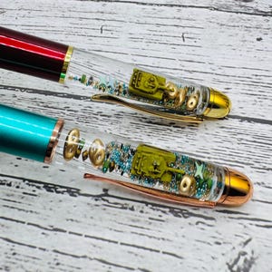 May include: Two pens with clear barrels, each containing a miniature olive-green tank, gold-colored accents, and small decorative elements. One pen has a burgundy top, the other is teal. Both have gold-colored tips and clips.