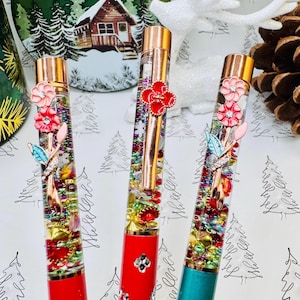 May include: Three decorative pens with clear barrels filled with colorful beads and glitter. The pens have red, red, and teal bases, and are topped with gold accents and floral embellishments. The background includes a decorative plate and a white reindeer.
