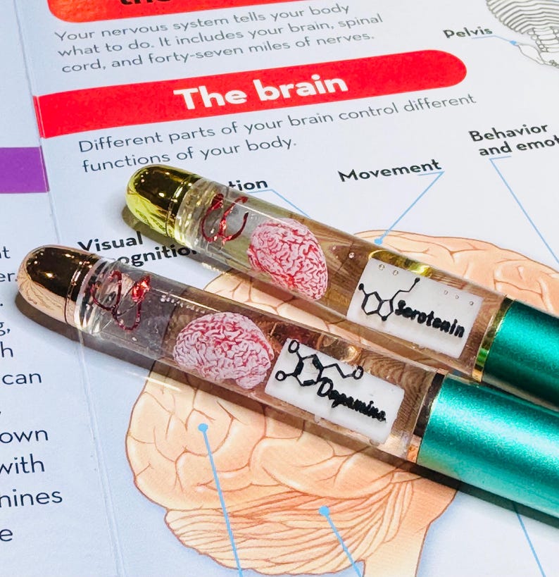Puede incluir: Dos bol&iacute;grafos transparentes con tapas doradas y detalles verde azulado. Cada bol&iacute;grafo contiene un modelo de cerebro en miniatura y una etiqueta con las palabras "Serotonina" y "Dopamina". Los bol&iacute;grafos se muestran en una p&aacute;gina de un libro sobre el cerebro.