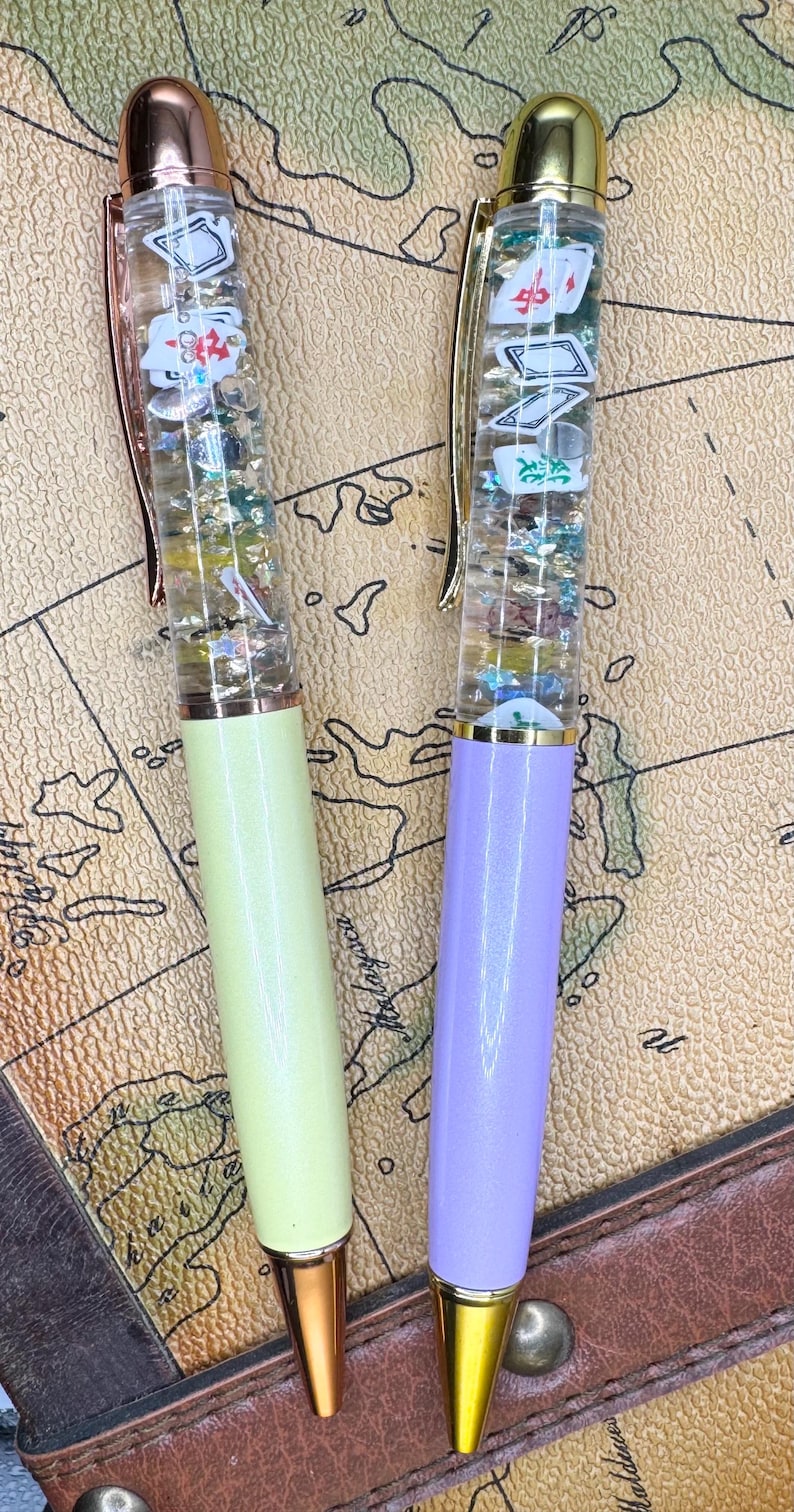 May include: Two clear pens with gold caps and gold accents. The pens are filled with glitter and small decorative items. One pen has a light green body and the other has a light purple body.