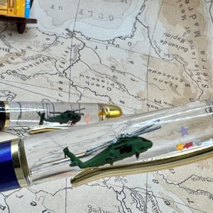 May include: Two pens with clear barrels containing miniature green helicopters and small colorful star-shaped floating pieces. One pen has a glossy blue cap and the other has a black cap, both featuring gold-toned clips and accents.