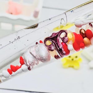 May include: A transparent glass tube filled with colourful beads and small sculptures. The tube contains red, white, and gold beads, along with a purple and silver anatomical sculpture. A yellow figure is visible in the background.