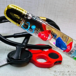 May include: A clear pen with a gold cap filled with small medical-themed charms, including stethoscopes and shoes. A black stethoscope and red scissors are in the foreground, suggesting a medical or nursing theme.