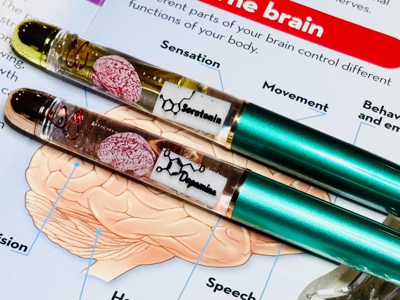 May include: Two clear glass tubes with gold caps filled with a liquid and a small brain model. The tubes are labeled 'Serotonin' and 'Dopamine' with their chemical structures. The tubes are on a white background with a diagram of the brain.
