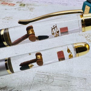 May include: Two fountain pens with clear barrels, each containing miniature law-themed elements. One pen has a black body with gold accents, the other is silver. Inside are tiny gavels, scales of justice, and books.