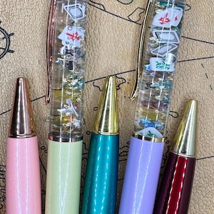 May include: Five colorful pens with gold-colored tips and accents. Two pens have clear barrels filled with floating Mahjong tiles and glitter. The other pens are solid colors: pink, green, teal, purple, and burgundy.