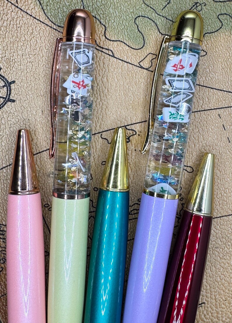 May include: Five colorful pens with gold-colored tips and accents. Two pens have clear barrels filled with floating Mahjong tiles and glitter. The other pens are solid colors: pink, green, teal, purple, and burgundy.