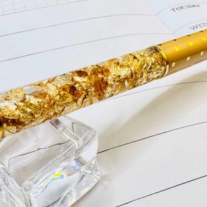 May include: A gold pen with gold leaf accents rests on a clear acrylic block, positioned over an open planner. The pen's barrel is filled with gold flakes, and the pen has a gold-colored tip. The planner displays the words "Weekly Plan" and days of the week.
