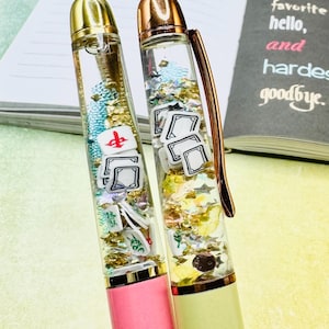 May include: Two decorative pens with clear barrels filled with glitter and small game-themed charms. One pen has a pink base, the other a light yellow base. The pens have gold-colored accents and are set against a notebook with text.