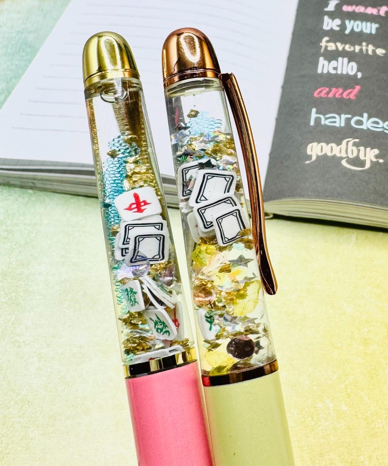 May include: Two decorative pens with clear barrels filled with glitter and small game-themed charms. One pen has a pink base, the other a light yellow base. The pens have gold-colored accents and are set against a notebook with text.