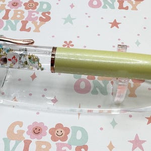 May include: A decorative pen with a clear barrel filled with small objects and glitter. The pen has a light green body, gold accents, and a rose gold clip. It rests on a clear plastic stand against a background with the text "GOOD VIBES ONLY".