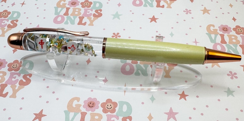 May include: A decorative pen with a clear barrel filled with small objects and glitter. The pen has a light green body, gold accents, and a rose gold clip. It rests on a clear plastic stand against a background with the text "GOOD VIBES ONLY".