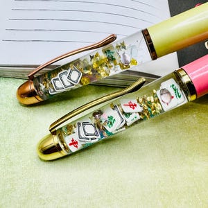May include: Two pens with clear barrels filled with gold flakes and miniature Mahjong tiles. One pen has a yellow barrel and a copper clip, the other has a pink barrel and a gold clip. Both pens have gold-colored tips.