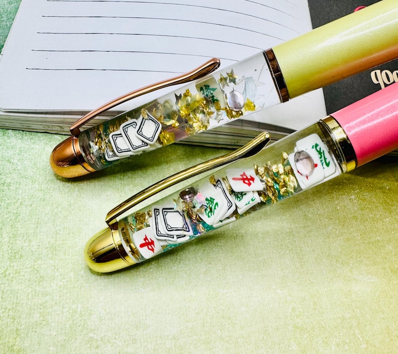 May include: Two pens with clear barrels filled with gold flakes and miniature Mahjong tiles. One pen has a yellow barrel and a copper clip, the other has a pink barrel and a gold clip. Both pens have gold-colored tips.