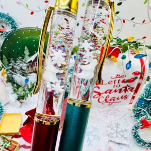 May include: Two Christmas-themed pens with gold detailing. One pen is burgundy, the other is teal. Both pens have a clear barrel filled with colourful glitter and a white snowman design. The pens are surrounded by Christmas decorations.