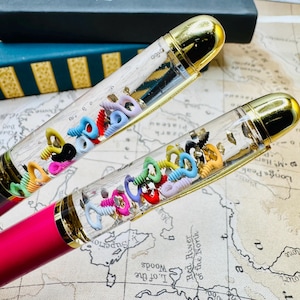 May include: Two fountain pens with clear barrels filled with colorful key-shaped charms and gold flakes. The pens have gold accents and pink and black bodies. The background includes a map and a book.