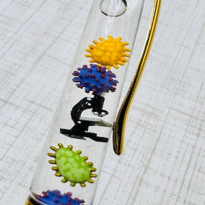 Könnte beinhalten: Ein transparenter Stift mit schwimmenden Anhängern, goldenem Verschluss und Clip. Im Inneren des durchsichtigen Schafts befinden sich Miniaturfiguren, darunter ein schwarzes Mikroskop, eine gelbe Zelle, eine grüne ovale Zelle und zwei blaue Formen.
