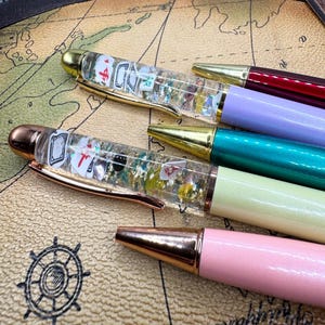 May include: A collection of colorful pens with gold and rose gold accents. Some pens have clear barrels filled with playing card-themed decorations and glitter. The pens are in shades of pink, green, purple, and burgundy.
