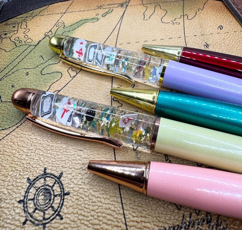 May include: A collection of colorful pens with gold and rose gold accents. Some pens have clear barrels filled with playing card-themed decorations and glitter. The pens are in shades of pink, green, purple, and burgundy.