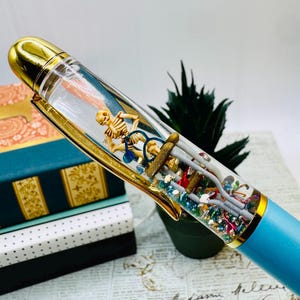 May include: A unique pen with a clear barrel containing a miniature skeleton riding a bicycle, medical tools, and colorful glitter. The pen has a gold-colored cap and clip, and a light blue barrel. The pen is a creative and whimsical writing instrument.