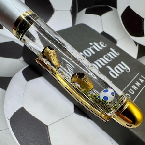 May include: A silver and gold pen with a clear barrel containing miniature gold soccer cleats, a soccer ball, and green accents. The pen rests on a journal with the text "Favorite Moment Day Journal".