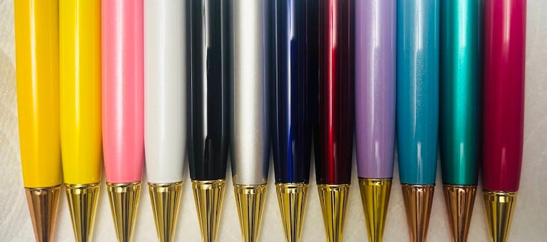 Puede incluir: Una colecci&oacute;n de bol&iacute;grafos coloridos con puntas doradas. Los bol&iacute;grafos est&aacute;n dispuestos en fila, mostrando una variedad de colores, incluyendo amarillo, rosa, blanco, negro, plateado, azul, rojo, morado, turquesa y magenta. Los bol&iacute;grafos tienen un acabado brillante.