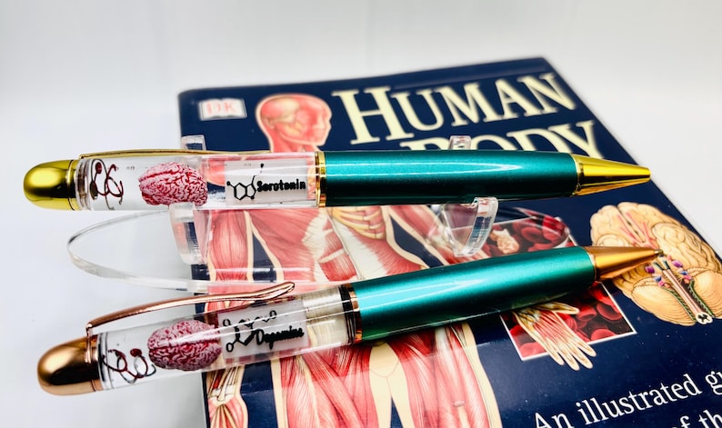 Puede incluir: Dos bol&iacute;grafos con barriles transparentes que contienen un modelo de cerebro y las palabras "Serotonina" y "Dopamina". Los bol&iacute;grafos tienen cuerpos de color verde azulado y detalles dorados. Se encuentran sobre un libro titulado "Human Body".