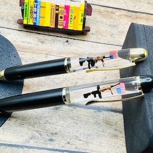 May include: Two black pens with gold accents and clear barrels. Inside each barrel is a miniature rifle and a small American flag. The pens are lying on a wooden surface.