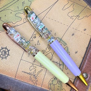 May include: Two decorative pens with clear barrels filled with small, colorful objects. One pen has a light green barrel and the other a lavender barrel. Both pens have gold and rose gold accents and are set against a map background.