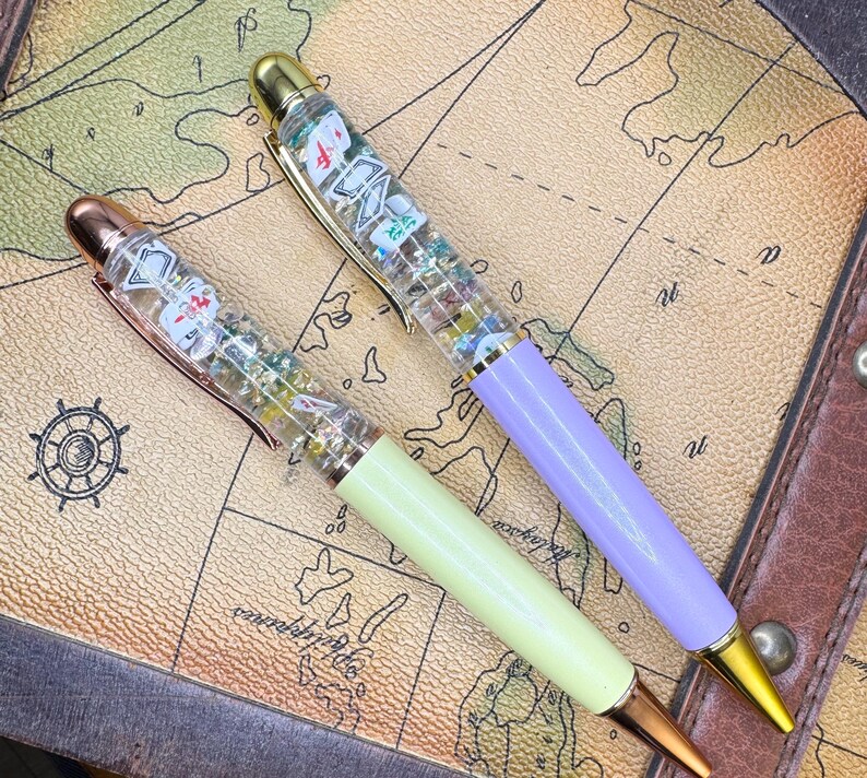 May include: Two decorative pens with clear barrels filled with small, colorful objects. One pen has a light green barrel and the other a lavender barrel. Both pens have gold and rose gold accents and are set against a map background.